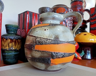 Puede incluir: Una colección de cerámica vintage en varios colores y patrones, incluyendo una jarra grande de color naranja y marrón con asa, un jarrón verde y marrón y un jarrón rectangular rojo y negro.