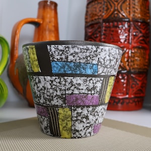 May include: A ceramic planter featuring a speckled black and white background with geometric rectangular accents in yellow, blue, and purple. The pot has a tapered shape and a dark rim, showcasing a textured surface pattern across the exterior.