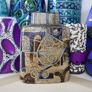 Puede incluir: Colección de jarrones de cerámica vintage de varias formas y tamaños. El jarrón central es rectangular con un diseño geométrico marrón y azul. Otros jarrones presentan patrones morados, azules y blancos. Los jarrones son probablemente artículos decorativos.