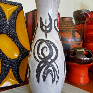 May include: A tall, light gray ceramic vase with a flared top and dark brown abstract designs. The vase features a textured surface and is surrounded by other decorative ceramic pieces in various colors and patterns.
