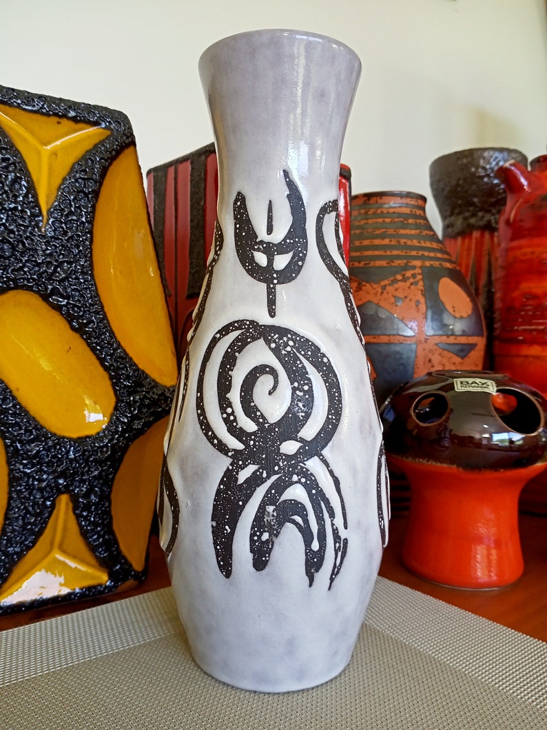 May include: A tall, light gray ceramic vase with a flared top and dark brown abstract designs. The vase features a textured surface and is surrounded by other decorative ceramic pieces in various colors and patterns.