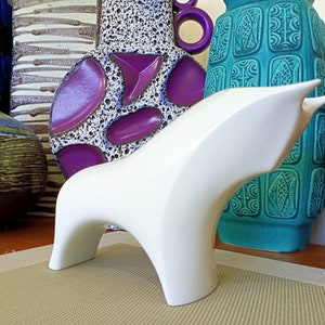 May include: White ceramic sculpture of a stylized bull, standing on a beige surface. The bull is positioned in front of a collection of colorful ceramic vases.