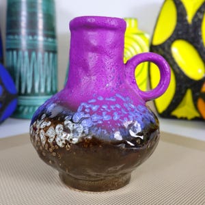 Puede incluir: Un pequeño jarrón de cerámica con asa, con una parte superior y un asa de color púrpura vibrante, que pasa a una base azul y marrón moteada. Otros jarrones coloridos son visibles en el fondo, lo que sugiere una colección de cerámica decorativa.