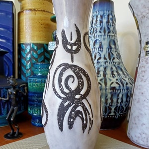 May include: A tall, light gray ceramic vase with dark brown abstract designs. The vase has a flared top and a textured surface. Other decorative vases are visible in the background.