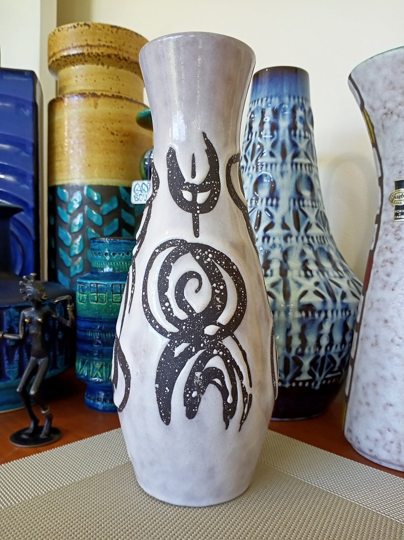 May include: A tall, light gray ceramic vase with dark brown abstract designs. The vase has a flared top and a textured surface. Other decorative vases are visible in the background.