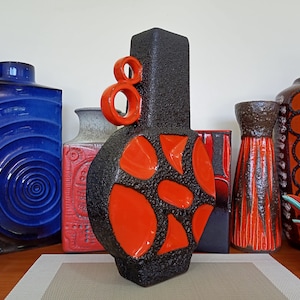 Puede incluir: Una colección de jarrones de cerámica de varias formas y colores. El jarrón central es negro con detalles en naranja y una superficie texturizada. Otros jarrones son azules, rojos y marrones, con patrones y diseños únicos. También se ve una figura decorativa de pájaro.