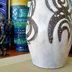 May include: A white ceramic vase with a textured surface and dark brown abstract designs. The vase has a wide base and a slightly narrower neck. Other decorative vases are visible in the background.