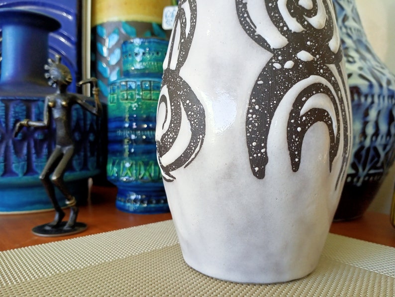 May include: A white ceramic vase with a textured surface and dark brown abstract designs. The vase has a wide base and a slightly narrower neck. Other decorative vases are visible in the background.