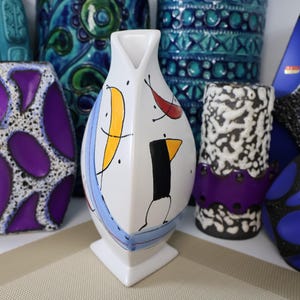 May include: A collection of ceramic vases in various shapes and sizes. The central vase is white with abstract yellow, black, and red painted designs. Other vases feature blue, purple, and black patterns. The vases are displayed on a neutral surface.