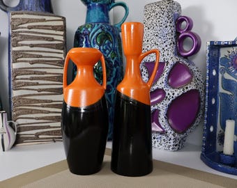 Puede incluir: Dos jarrones de cerámica con diseño bicolor que presenta una sección superior naranja brillante y una sección inferior negra sólida. El jarrón izquierdo tiene dos asas pequeñas, mientras que el derecho tiene una sola asa y cuello esbelto.