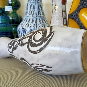 May include: A white ceramic vase with a wide base and a narrow neck, decorated with black abstract swirls. The vase is lying on its side, showcasing its shape and design. Other decorative vases are visible in the background.
