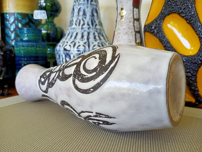 May include: A white ceramic vase with a wide base and a narrow neck, decorated with black abstract swirls. The vase is lying on its side, showcasing its shape and design. Other decorative vases are visible in the background.