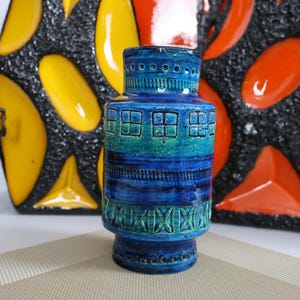 May include: A blue ceramic vase with a textured surface and geometric patterns. The vase features shades of blue and teal, with a cylindrical shape and a wide base. Decorative vases in the background are yellow and orange.