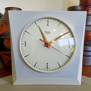May include: A white square clock with black hands and a red second hand. The clock face is white with black numbers and a gold 'Dufa electro' logo. The clock is sitting on a wooden surface.