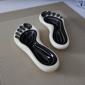 May include: Two ceramic dishes shaped like human footprints, featuring a glossy cream-colored border and a deep black interior basin. The black area is decorated with white concentric circles on the toes and abstract lines on the sole.