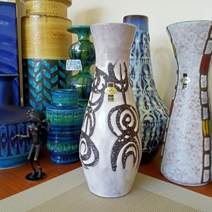 May include: A collection of vintage ceramic vases in various shapes, sizes, and colors. The central vase is light gray with a black abstract design. Other vases are blue, green, and brown, with textured and patterned surfaces. A small bronze figurine is in the foreground.