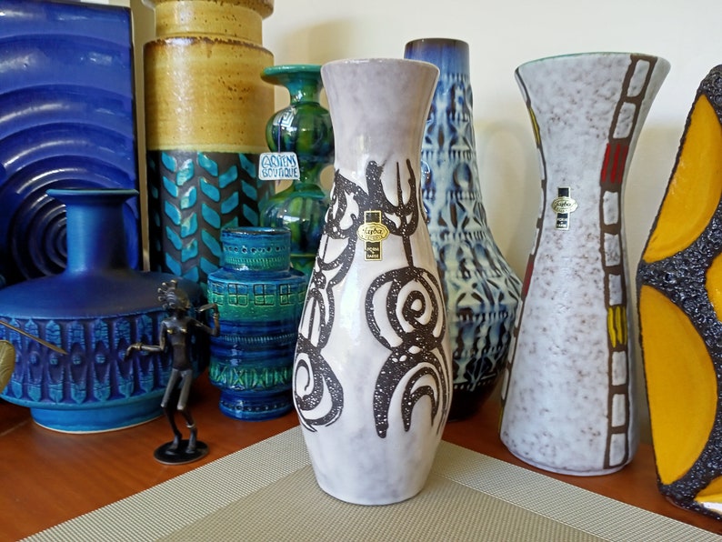 May include: A collection of vintage ceramic vases in various shapes, sizes, and colors. The central vase is light gray with a black abstract design. Other vases are blue, green, and brown, with textured and patterned surfaces. A small bronze figurine is in the foreground.