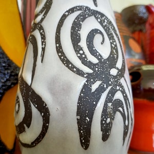 May include: A white ceramic vase with a black spiral design and speckled texture. The vase is part of a collection of decorative ceramic pieces, including a yellow and black butterfly-shaped vase and other colorful items.
