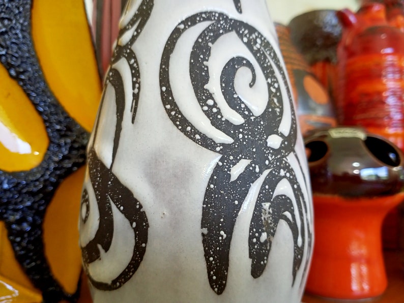May include: A white ceramic vase with a black spiral design and speckled texture. The vase is part of a collection of decorative ceramic pieces, including a yellow and black butterfly-shaped vase and other colorful items.