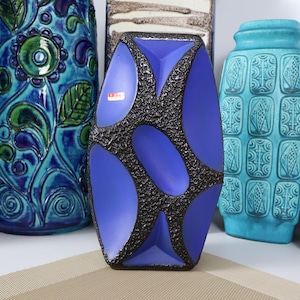 May include: A tall, rectangular ceramic vase featuring a vibrant blue smooth finish contrasted by a dark, textured, crater-like pattern. The vase has a small rectangular sticker on the front and is surrounded by other colorful ceramic vessels.