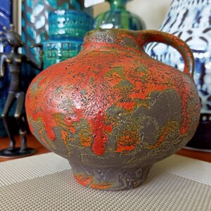 Può includere: Un vaso in ceramica marrone con una glassa rossa e arancione. Il vaso ha un corpo rotondo e un collo corto con un manico.