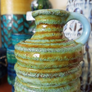 May include: A small, green and brown ceramic pitcher with a rounded body and a handle. The pitcher has a textured surface with a glossy finish.