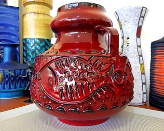 May include: A red ceramic vase with a stylized fish design. The vase has a wide base and a narrow neck, with a handle on one side. The fish design is embossed on the vase and is painted in black.