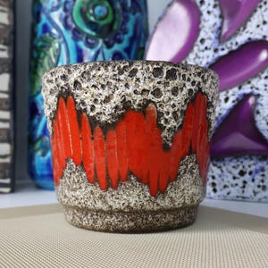 May include: A ceramic planter featuring a textured, speckled off-white and dark brown base with a vibrant, glossy red band encircling the middle. The rim and lower section exhibit a porous, crater-like surface texture, contrasting with the smooth red glaze.