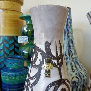 May include: A collection of vintage ceramic vases in various shapes and colors. The foreground features a tall, light gray vase with a black abstract design and a "Jasba Keramik" label. Other vases include blue, green, and brown tones.