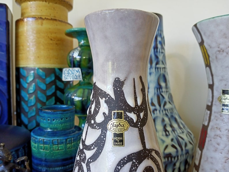 May include: A collection of vintage ceramic vases in various shapes and colors. The foreground features a tall, light gray vase with a black abstract design and a "Jasba Keramik" label. Other vases include blue, green, and brown tones.