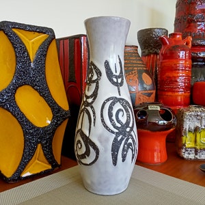 May include: A collection of ceramic vases in various shapes and sizes. The vases feature a range of colors, including orange, black, yellow, and white. One tall, slender vase has a gray base with black abstract designs. The vases are displayed on a wooden shelf.