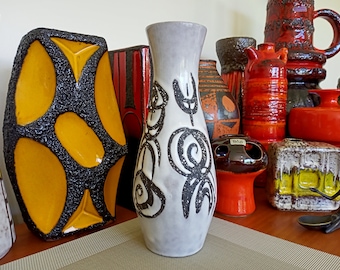May include: A collection of ceramic vases in various shapes and sizes. The vases feature a range of colors, including orange, black, yellow, and white. One tall, slender vase has a gray base with black abstract designs. The vases are displayed on a wooden shelf.
