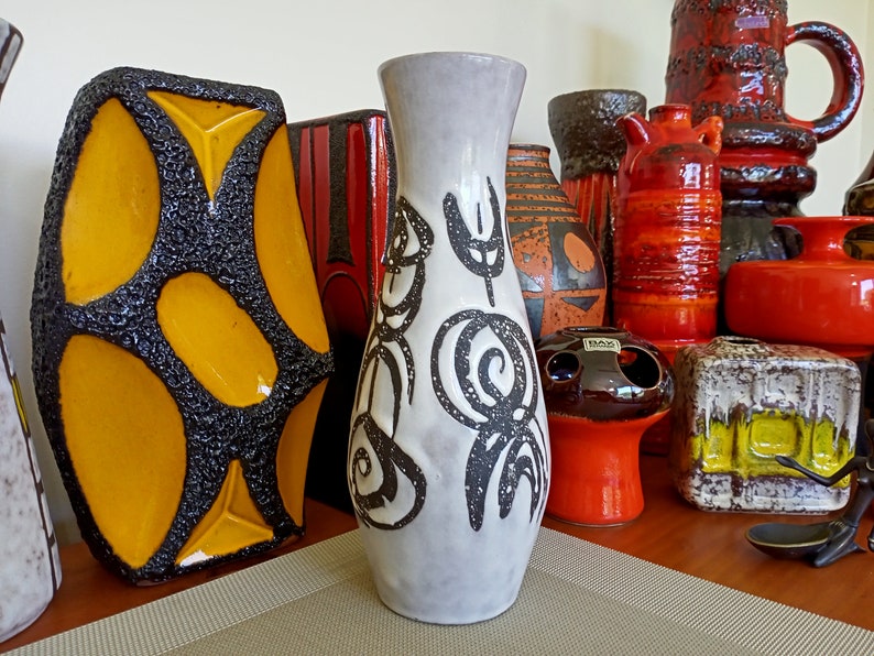 May include: A collection of ceramic vases in various shapes and sizes. The vases feature a range of colors, including orange, black, yellow, and white. One tall, slender vase has a gray base with black abstract designs. The vases are displayed on a wooden shelf.