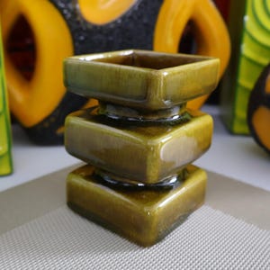 Puede incluir: Un jarrón de cerámica verde oliva apilado, con una parte superior e inferior cuadradas. El jarrón tiene un acabado brillante y está rodeado de otros jarrones decorativos en tonos amarillos, negros y verdes. El jarrón mide unos 10 cm de alto.