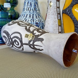 May include: A white ceramic vase with a brown interior and a decorative black swirl pattern. The vase has a label that reads "Harba Paris". Other decorative vases are visible in the background.