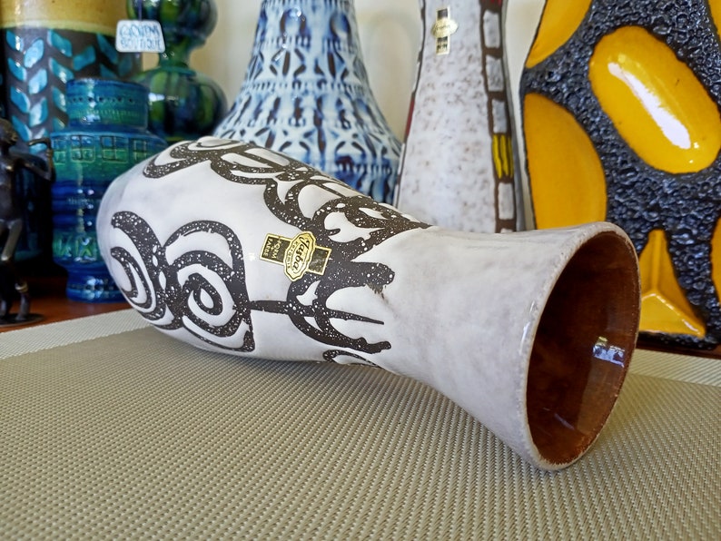 May include: A white ceramic vase with a brown interior and a decorative black swirl pattern. The vase has a label that reads "Harba Paris". Other decorative vases are visible in the background.