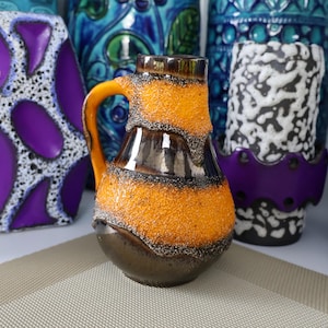 Puede incluir: Una jarra de cerámica con base marrón, parte media texturizada naranja y parte superior marrón. El asa es naranja. Otras coloridas jarrones están en el fondo, incluyendo diseños morados, azules y blancos. La jarra es un artículo decorativo.