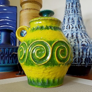 Puede incluir: Una jarra de cerámica amarilla con un diseño de espiral verde. La jarra tiene un asa y un cuerpo redondeado.