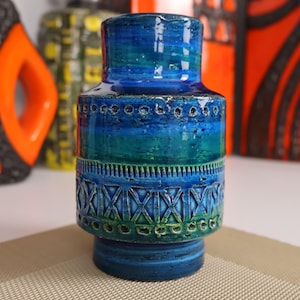 May include: A ceramic vase in shades of blue and green. The vase has a cylindrical form with a flared base and a narrower neck. The surface is decorated with textured patterns and a glossy finish, typical of mid-century design.
