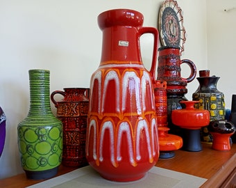 Może przedstawiać: Kolekcja vintage ceramicznych wazonów w różnych kolorach i wzorach. Największy wazon jest czerwony z białymi i pomarańczowymi akcentami i ma uchwyt. Pozostałe wazony są zielone, brązowe i czarne z geometrycznymi wzorami.