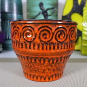 Puede incluir: Una maceta de cerámica naranja con un diseño de espirales y líneas verticales negras. La maceta tiene una parte superior ancha y una base cónica. Otros jarrones decorativos están en el fondo.