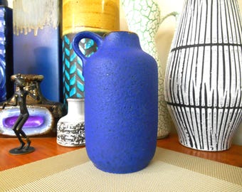 May include: A blue ceramic vase with a textured surface and a handle. The vase is sitting on a table with other vases.