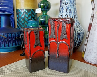 May include: Two rectangular ceramic vases. The vases are red with dark brown, textured, dripped designs. The bottom of the vases are dark brown. Other ceramic vases are in the background.