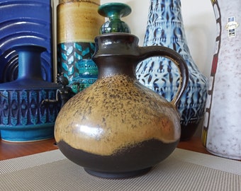May include: A brown and tan ceramic pitcher with a speckled glaze and a handle. The pitcher has a rounded body and a wide mouth.