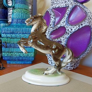 May include: A brown ceramic horse figurine with a white base. The horse is standing on its hind legs with its front legs raised. The figurine is on a beige surface with a purple and white ceramic vase in the background.