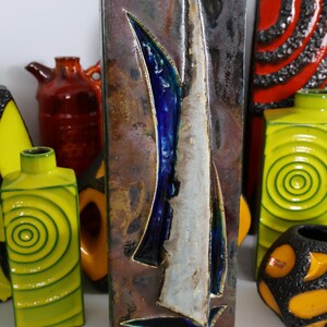 Może przedstawiać: Prostokątny ceramiczny wazon z motywem żaglówki. Wazon ma brązową, niebieską i białą kolorystykę. Inne kolorowe wazony znajdują się w tle.