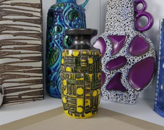 May include: A cylindrical ceramic vase featuring a vibrant yellow and dark green geometric relief pattern. The vase has a smooth, dark brown neck and rim, contrasting with the textured, blocky design that covers the main body of the vessel.