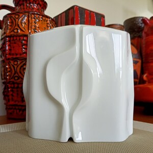 May include: A white ceramic vase with a modern, abstract design. The vase has a smooth, glossy finish and features a series of curved lines and shapes.