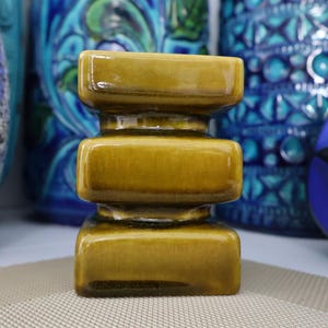 May include: A small, olive-green ceramic vase with a stacked rectangular design. The vase has a glossy finish and is surrounded by other decorative vases in shades of blue and purple.