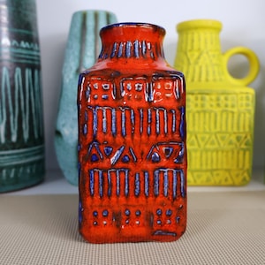 Könnte beinhalten: Eine rechteckige, orangefarbene Keramikvase mit strukturierter Oberfläche und blauen Akzenten. Die Vase weist ein geometrisches Muster auf. Im Hintergrund sind weitere farbige Vasen zu sehen, die eine Sammlung dekorativer Keramik zeigen.